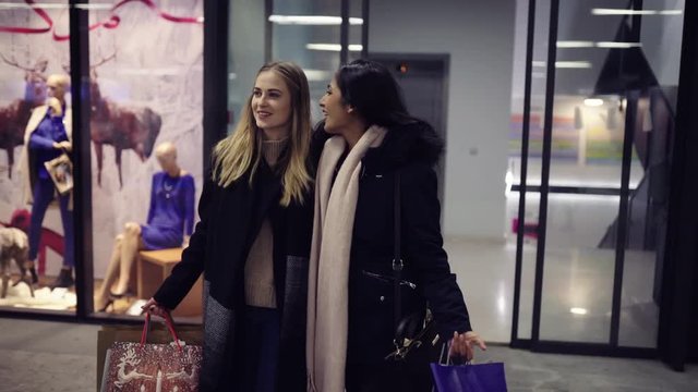 Two cute young adolescent women, heavy loaded with christmas gifts,  shopping, while walking along store windows and choosing and admiring the items. 