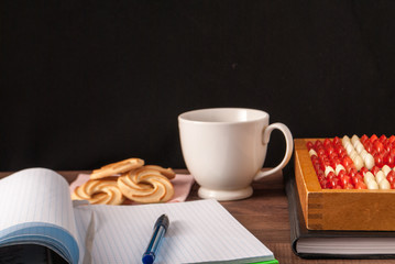 abacus on a wooden surface and a notebook with a ballpoint pen and a white cup with biscuits