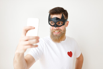 Waist up portrait of cheerful bearded male model poses at smart phone camera, wears batman mask and...