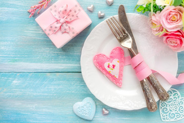 Romantic dinner concept. Table place setting with a white plate, vintage silverware tied with a pink ribbon and many different heart shape decorations, a gift and flowers