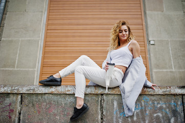 Stylish curly blonde model girl wear on white with cup of coffee posing against shutter background.
