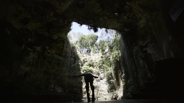 from below big natural cave with lians, trees and waterfals on the walls,  young men walking and jumping off cliff to water
