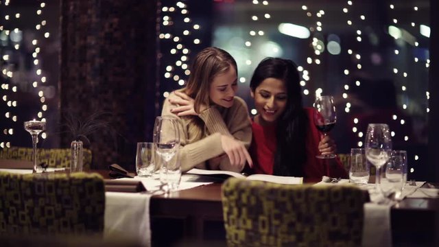 Two adolescent women going through the menue  in a fancy restaurant, while drinking a glass of red wine and laughing and talking, maybe gossiping