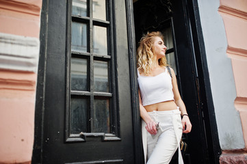 Stylish curly blonde model girl wear on white posing against old wooden door.