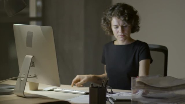 Tracking shot of attractive businesswoman sitting at office desk at night, reading documents and working on computer