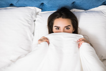 . Cheerful girl in bed hiding her face under the blanket. Teenage girl lying under a blanket. Woman playful with a blanket view from above. Girl peeks out from the duvet.