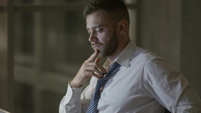 Young tired businessman loosening his tie while sitting at work in the evening and reading documents