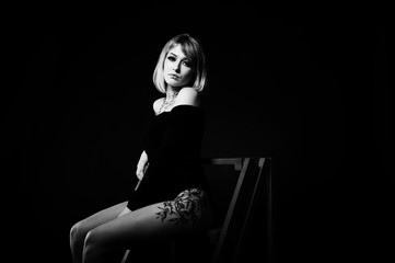 Obraz na płótnie Canvas Studio portrait of blonde girl with originally make up on neck and tattoo on thigh, wear on black dress at dark background, sitting on red ladder.