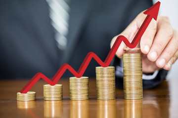 Businessman hand holding one coin and red arrow up on money coin stack arranged as a graph on...