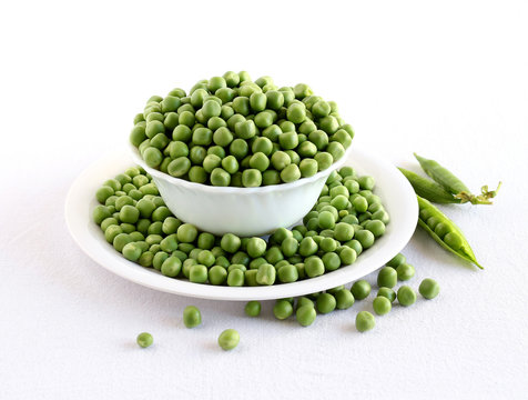 Peas, fresh and organic, in a bowl on a plate with spilled peas, and in the background are pea pods.