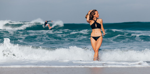 Beautiful girl in black swimwear coming out of the ocean with surfer riding the wave in the background