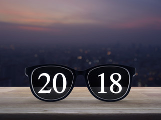2018 white text with black eye glasses on wooden table over blur aerial view of city tower on warm light sundown, Happy new year business concept