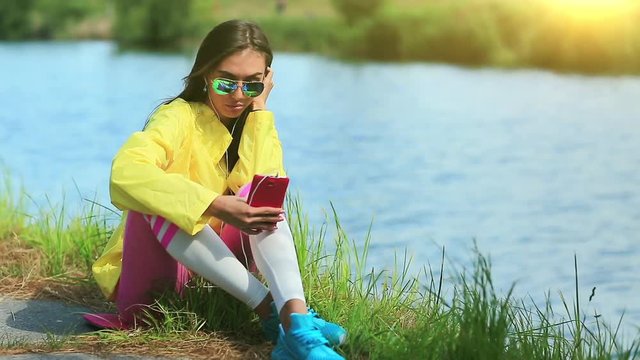 Sportswoman communicates via smartphone. Girl sits near lake and talks on smartphone. Woman in sportswear with smartphone communicates through skype or videochat, beautiful sun at background