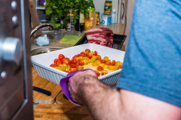 Male hand holding white ceramic baking tray with chopped tomatoes over raw beef roast kitchen table