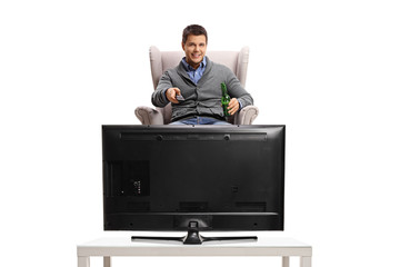 Young guy with a remote control and a bottle of beer sitting in an armchair and watching television