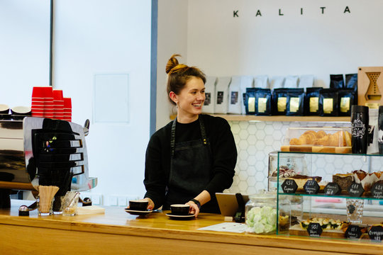 Friendly hospitable woman barista holding a two coffee cups with smiling face at cafe counter background, food and drink industry concept. Modern coffee shop interior.