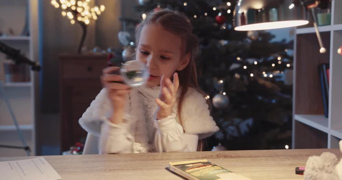Cute girl sit on the table smile hold small toy minature near Christmas tree in home figurine globe holidays joy kids lights child holiday happy tradition winter slow motion
