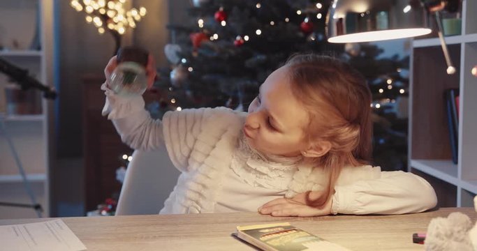 Smiling girl sit on the table smile hold small toy minature near Christmas tree in home figurine globe holidays joy kids lights child holiday happy portrait close up tradition winter slow motion