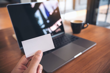 A hand holding white empty business card with laptop and coffee cup on wooden table in office