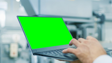 Close-up of the Engineer Holding Laptop with Green Screen Chroma Key Template Great for Mockup. In the Background Modern Factory Equipment.