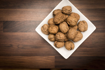 A bowl of whole walnuts in their shells in a white square bowl shot from above on a black slate background