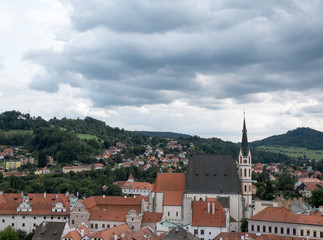 Obraz premium View of the charming medieval town, Cesky Krumlov, from the castle, view of St Vitus Chruch (Kostel vs Vitas) and the red roofed village surrounding the church. Stormy clouds and green hills.