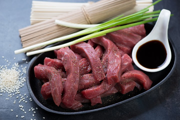 Cast-iron pan with fresh uncooked beef meat slices, soy sauce, green onion and noodles, studio shot
