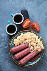 Scrambled eggs with sausages and coffee for breakfast. Flat-lay on a blue stone background, vertical shot