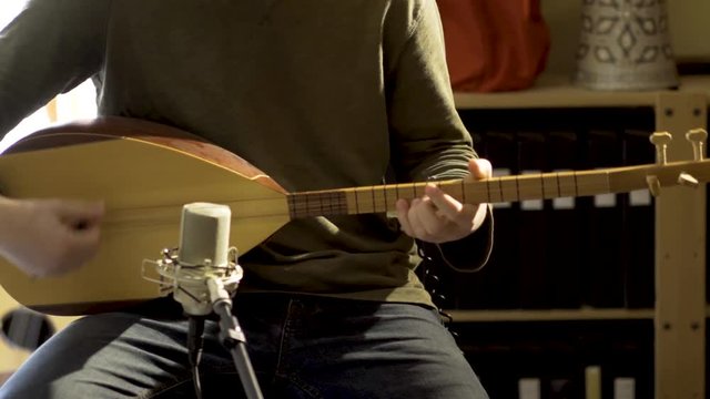 Tight shot slide to the left of young man playing baglama saz with microphones in front in a home studio.