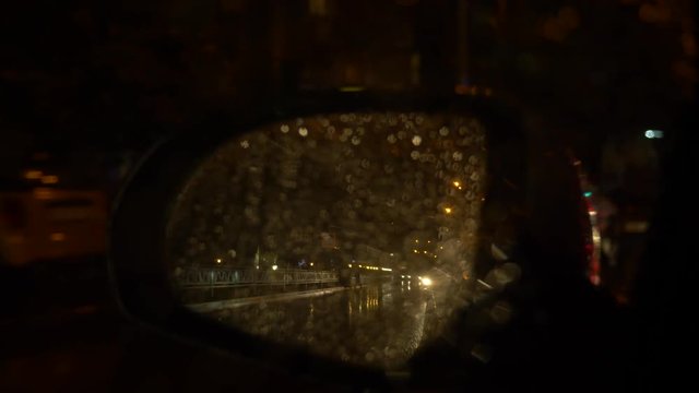 rainwater flow on the rearview mirror. The car stops next to the road with beautiful colorful blurry traffic lights off the road. 4k