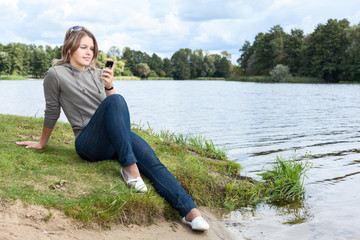 Blond woman sitting on river bank with mobile phone in hand, looking at screen