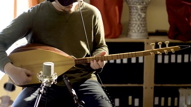 Static shot of young man wearing headphones playing baglama saz with microphones in front of instrument and oud and darbuka drums in background in a room with a lot of light and camera slide to the ri