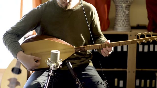 Very slow slide of young man wearing headphones playing baglama saz with microphones in front of instrument and oud and darbuka drums in background in a room with a lot of light and camera slide to th