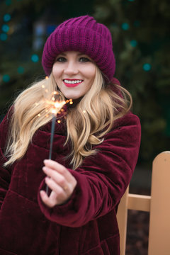 Closeup shot of adorable blonde woman dressed in warm clothes, holding glowing sparklers at the Christmas tree