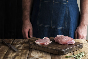 a man in a kitchen apron, meat on a board and a wooden table, rosemary and a knife