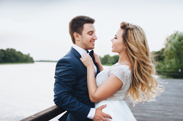 Beautiful and young bride and groom tenderly embrace on the pier by the river. Attractive bride with long blond hair. Wedding ceremony in nature by the river.