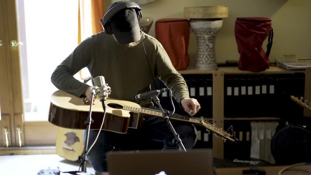 Wide camera slide to left as young man wearing headphones and playing slide guitar with microphones and computer in home recording studio.