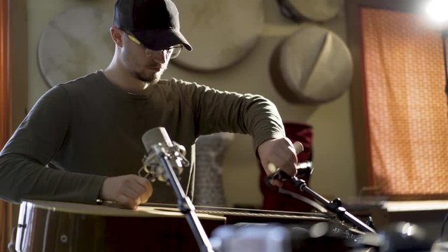 Young man with ball cap adjusting microphones preparing to play music with a slide guitar.  Doumbek and frame drums in the background.