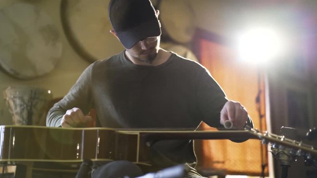 Medium tight shot of young man playing baglama saz with microphones in front of instrument and oud in corner in a room with a lot of light.