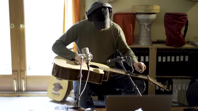 Wide camera slide to right as young man wearing headphones and playing slide guitar with microphones and computer in home recording studio.