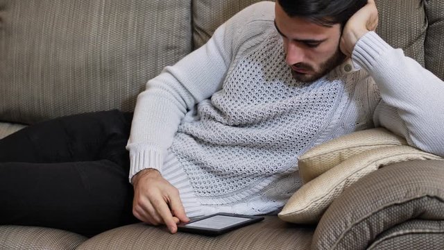 Handsome young man at home smiling, reading with ebook reader lying on a couch