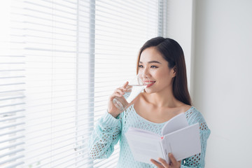 Beautiful asian girl drinking water and reading book near window at home