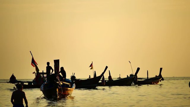 Group of silhouette long tail boat converted to boat excursions floating in the andaman sea with golden light of the Sun  before sunset and yatch boat background in travel or transportation concept.