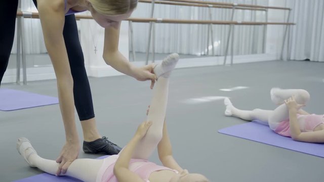 Female school coach keeps foot in correct position of girl on mat. During ballet lessons coach helps pupils stretch limb muscles.