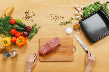 Raw meat. Fresh beef tenderloin on cutting board on wooden table with different vegetables, spices, mushrooms, baking tray, knife. Top view flat lay Copy space for advertisement. Woman cuts with knife