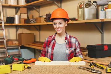 Beautiful caucasian young brown-hair woman in plaid shirt, gray T-shirt, yellow gloves, protective helmet working in carpentry workshop at wooden table place with piece of wood, different tools.