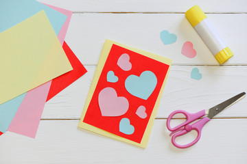 Valentines day or Mothers day card with pink and blue hearts, scissors, glue stick, colored paper sheets on a wooden table. Easy art and craft with paper for kids. Top view