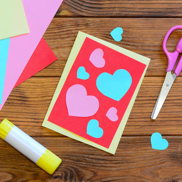 Valentines day or Mothers day greeting card with pink and blue hearts. Scissors, glue stick, colored paper sheets on a wooden table. Easy paper craft work idea for kids. Top view