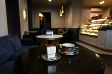 A horizontal image of a tiramisu dessert in a transparent glass and a blue cup of coffee with a biscuit near by. Wooden table in a cafe.