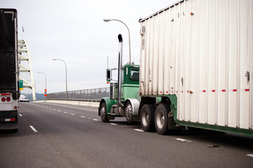 Big rig green semi truck with bulk trailer going on bridge beside another semi truck